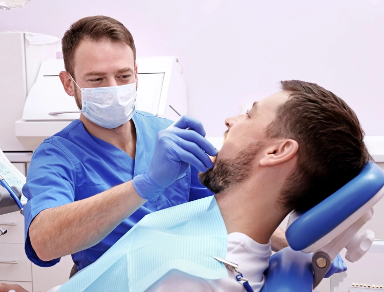 Dentist wearing blue scrubs and mask examining a male patient's teeth in a dental chair.